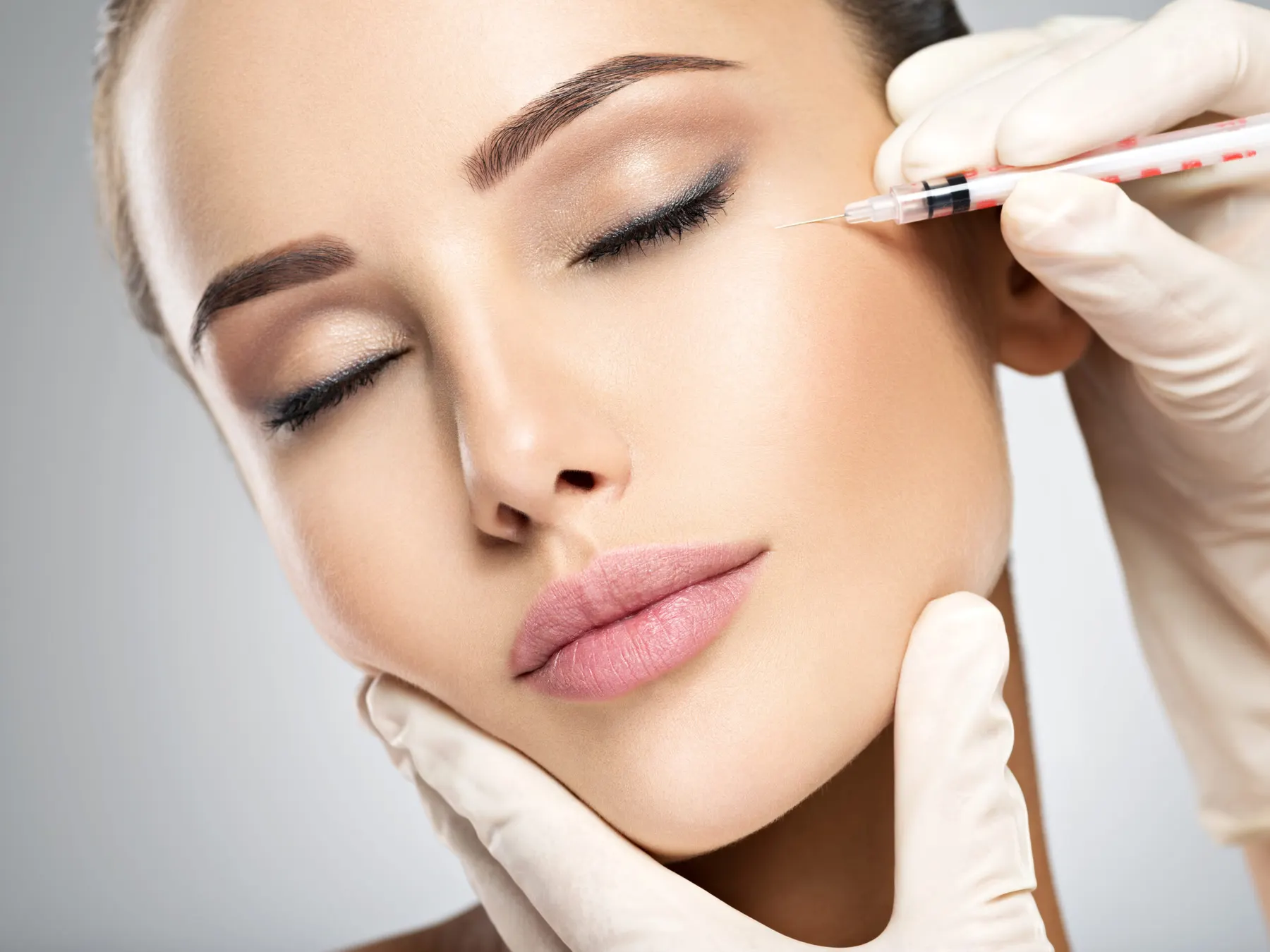 A close-up of a client with her eyes closed while white-gloved hands hold a fine syringe beside her brow.