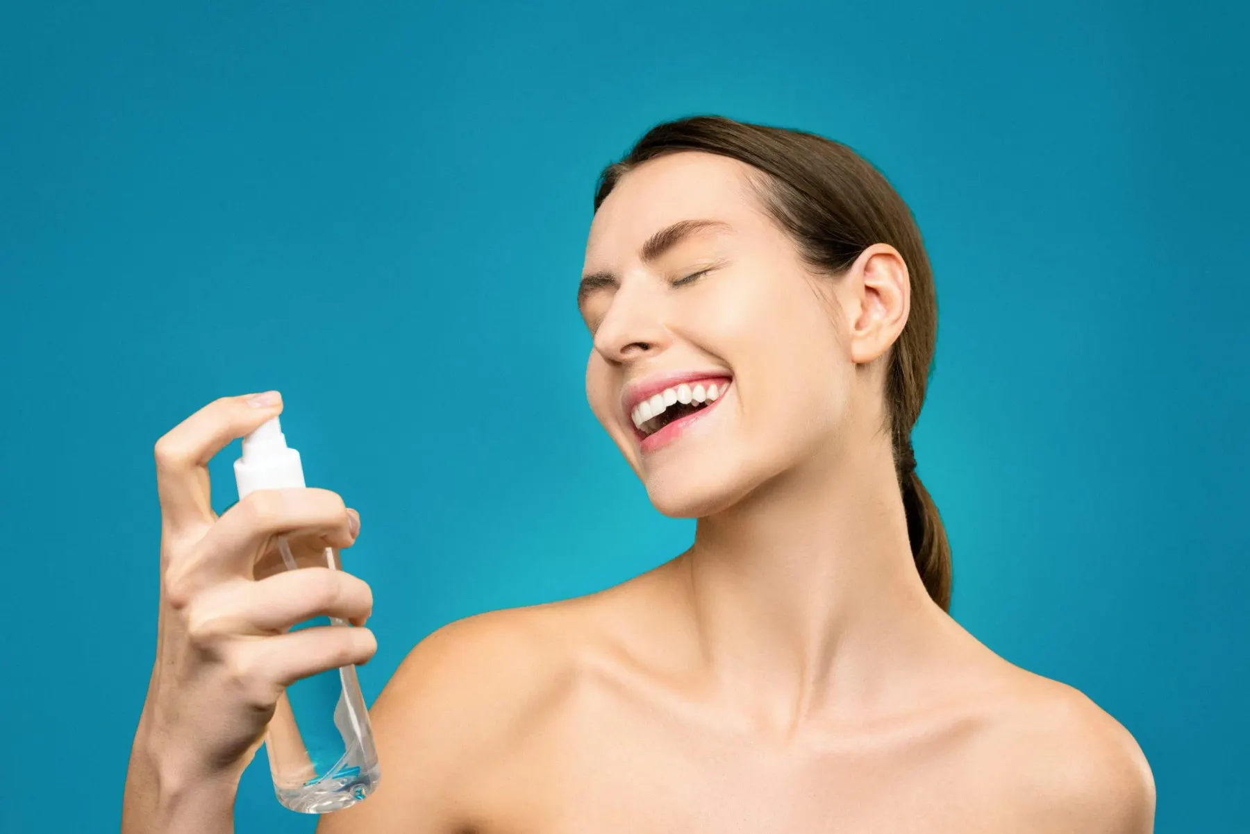 A woman laughing while spraying a facial mist, photographed against a teal background