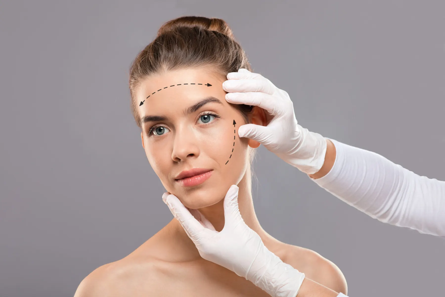 A woman looking at the camera while gloved hands hold a measuring guide against her cheek and dotted planning lines run across her face.