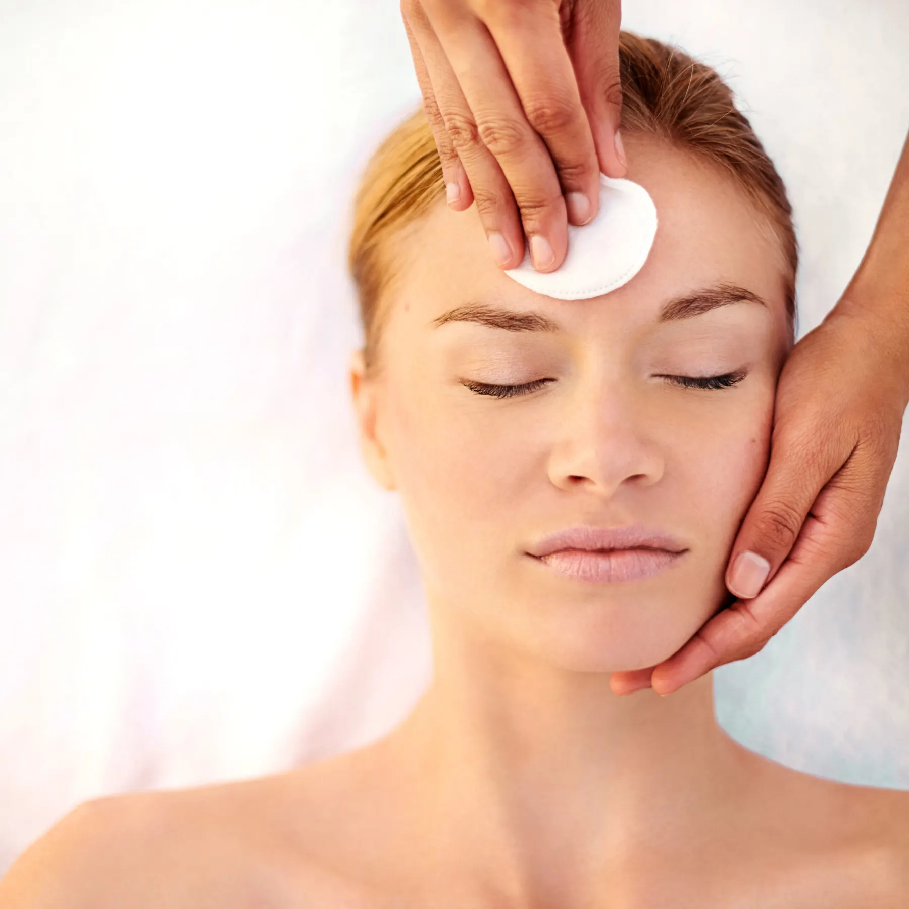 Two hands holding a cotton pad against the forehead of a client resting on a white towel.